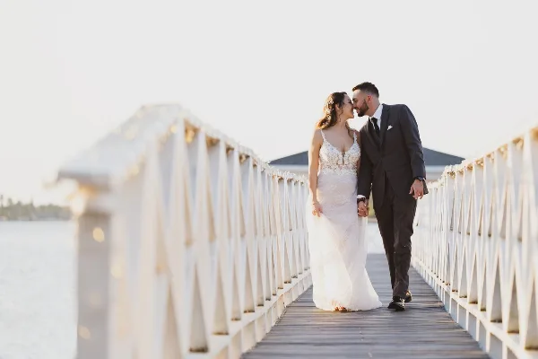 A bride and groom standing on a wooden bridge over water, holding hands and looking at each other.