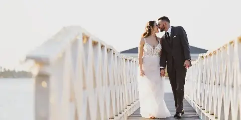 A bride and groom standing on a wooden bridge over water, holding hands and looking at each other.