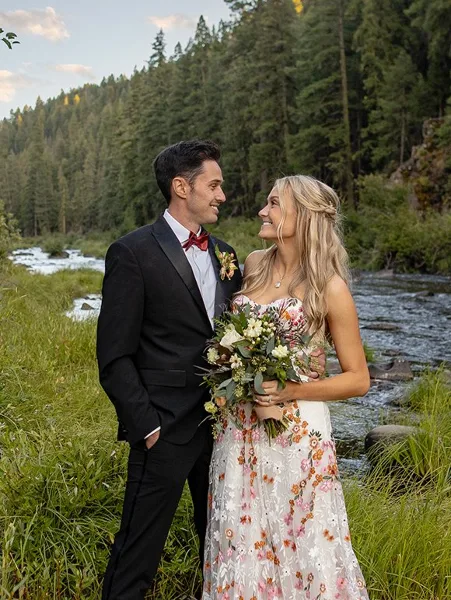 A bride and groom standing together in a forest clearing next to a flowing river.