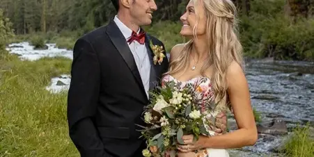 A bride and groom standing together in a forest clearing next to a flowing river.