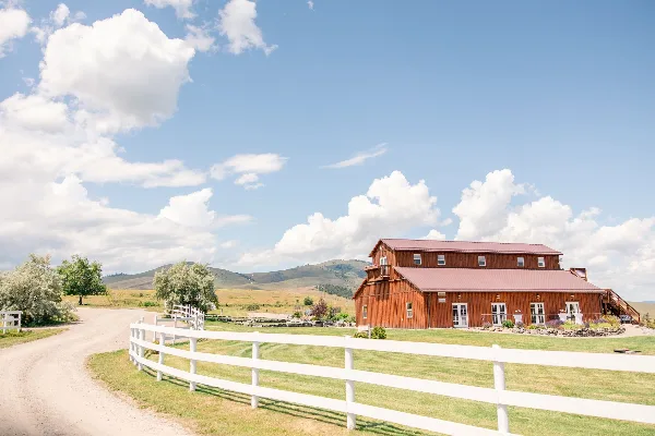 A large wooden barn with a red roof and white fencing situated in a grassy field with mountains in the background.