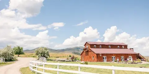 A large wooden barn with a red roof and white fencing situated in a grassy field with mountains in the background.