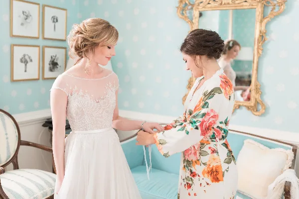 A bride with a romantic updo hairstyle and natural makeup standing in a room with light blue wallpaper, being assisted by another woman.