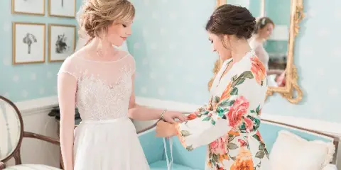 A bride with a romantic updo hairstyle and natural makeup standing in a room with light blue wallpaper, being assisted by another woman.