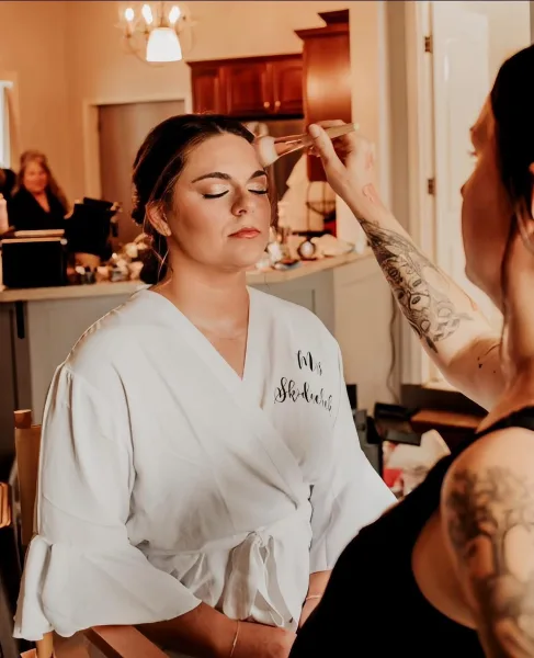 A makeup artist applying makeup to a bride in a white robe.