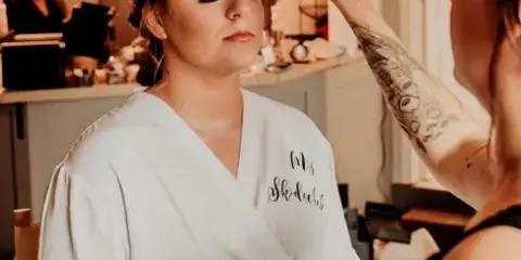 A makeup artist applying makeup to a bride in a white robe.