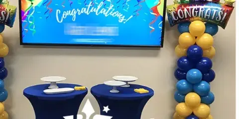 A corporate event space decorated with blue and yellow balloon columns, a 'Congratulations' balloon, and a table with a Blank Canvas Event Decor skirt.
