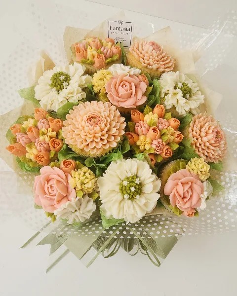 A bouquet of cupcakes decorated with hand-piped buttercream flowers in shades of pink, white, and yellow, arranged with green leaves and wrapped in cellophane.