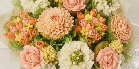 A bouquet of cupcakes decorated with hand-piped buttercream flowers in shades of pink, white, and yellow, arranged with green leaves and wrapped in cellophane.