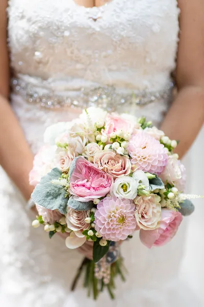 A close-up of a bride in a white lace dress holding a large, round bouquet of pink and white roses, dahlias, and greenery.