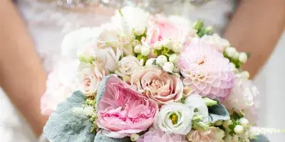 A close-up of a bride in a white lace dress holding a large, round bouquet of pink and white roses, dahlias, and greenery.