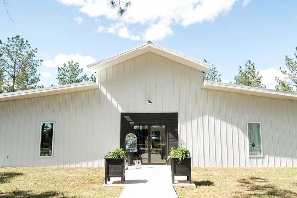 Exterior view of a modern white metal event venue building with a dark entryway, surrounded by pine trees under a blue sky.