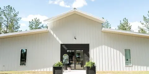 Exterior view of a modern white metal event venue building with a dark entryway, surrounded by pine trees under a blue sky.
