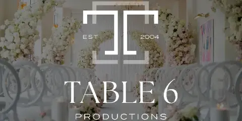 A luxury wedding ceremony aisle featuring white chairs, candles, and large floral arrangements with a Table 6 Productions logo overlay.