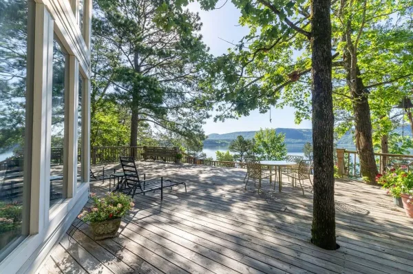 A spacious wooden deck with outdoor furniture and potted plants, attached to a white house, overlooking a lake and forested hills.