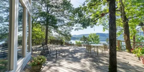 A spacious wooden deck with outdoor furniture and potted plants, attached to a white house, overlooking a lake and forested hills.
