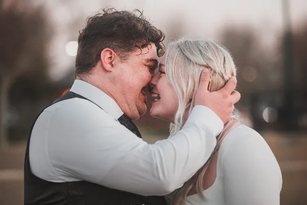 A candid, close-up photograph of a bride and groom laughing together outdoors, with the groom wearing a white shirt and dark vest and the bride in a white top.
