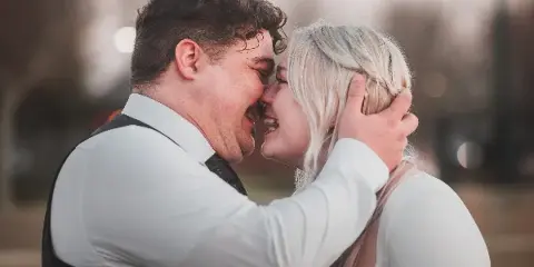 A candid, close-up photograph of a bride and groom laughing together outdoors, with the groom wearing a white shirt and dark vest and the bride in a white top.
