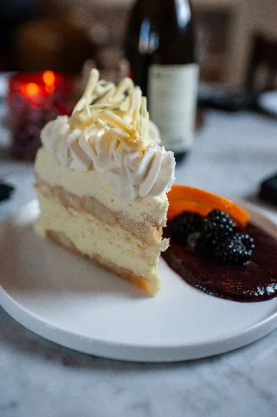 A slice of layered cream cake topped with white frosting and chocolate shavings, served on a white plate with a side of berry compote.