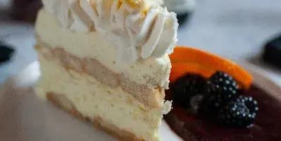 A slice of layered cream cake topped with white frosting and chocolate shavings, served on a white plate with a side of berry compote.
