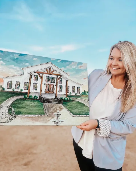 A woman holding a painting of a white wedding venue building with a fountain in the foreground.