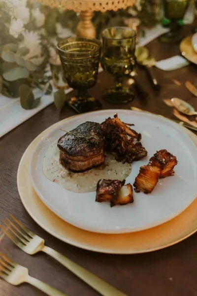 A plated meal with a seared protein and roasted vegetables served on a white plate with gold-rimmed charger plates and vintage glassware.