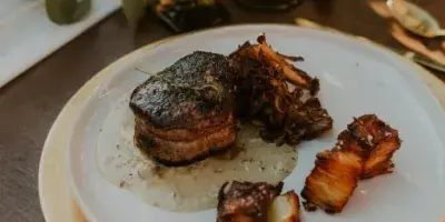 A plated meal with a seared protein and roasted vegetables served on a white plate with gold-rimmed charger plates and vintage glassware.