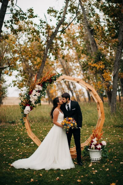 A bride and groom sharing a kiss under a rustic wooden arch decorated with flowers and greenery in an outdoor field setting.