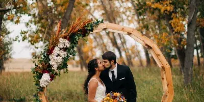 A bride and groom sharing a kiss under a rustic wooden arch decorated with flowers and greenery in an outdoor field setting.