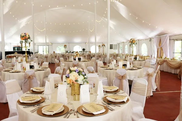 Decorated banquet tent interior with round tables, white linens, gold charger plates, place settings, and floral centerpieces.