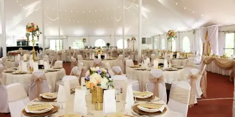 Decorated banquet tent interior with round tables, white linens, gold charger plates, place settings, and floral centerpieces.
