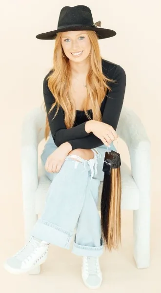 A woman with long red hair wearing a black hat sits in a chair holding a packaged hair extension.