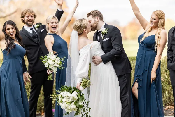 A bride and groom share a kiss surrounded by their smiling wedding party in an outdoor setting.