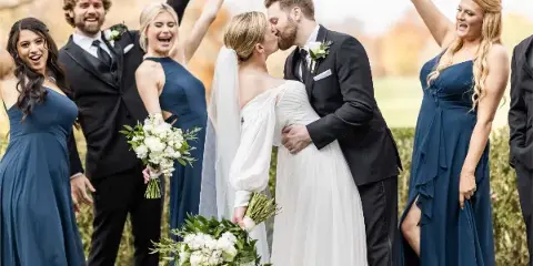 A bride and groom share a kiss surrounded by their smiling wedding party in an outdoor setting.