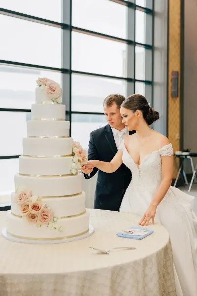 A bride and groom cutting a tall, white, multi-tiered wedding cake decorated with pink roses and gold bands.