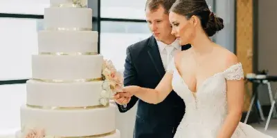 A bride and groom cutting a tall, white, multi-tiered wedding cake decorated with pink roses and gold bands.