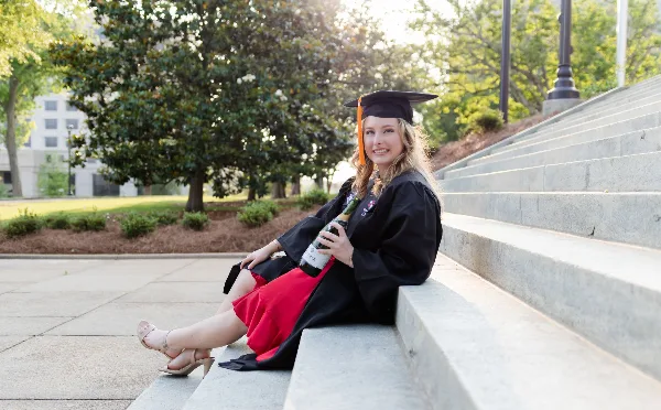 A young woman in a graduation cap and gown sitting on concrete steps holding a bottle of champagne.