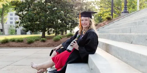 A young woman in a graduation cap and gown sitting on concrete steps holding a bottle of champagne.