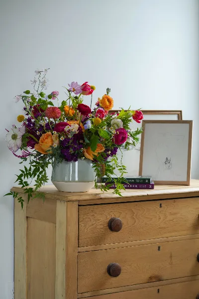 A colorful, garden-style floral arrangement in a light blue ceramic vase sitting on a wooden dresser.