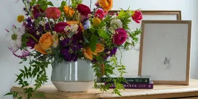 A colorful, garden-style floral arrangement in a light blue ceramic vase sitting on a wooden dresser.