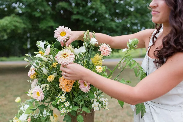 A person arranging a wedding bouquet with pink daisies, white flowers, and greenery outdoors.