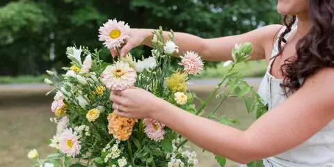 A person arranging a wedding bouquet with pink daisies, white flowers, and greenery outdoors.