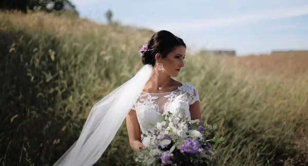 A bride in a white lace wedding dress and veil stands in a field of tall grass, holding a bouquet of white and purple flowers.
