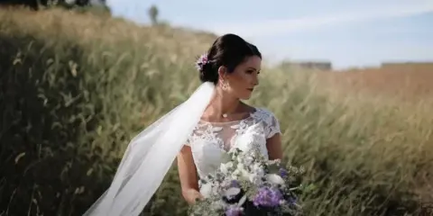 A bride in a white lace wedding dress and veil stands in a field of tall grass, holding a bouquet of white and purple flowers.