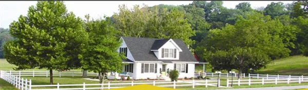 White farmhouse with dark roof on a green farm property surrounded by trees and white fencing