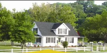 White farmhouse with dark roof on a green farm property surrounded by trees and white fencing