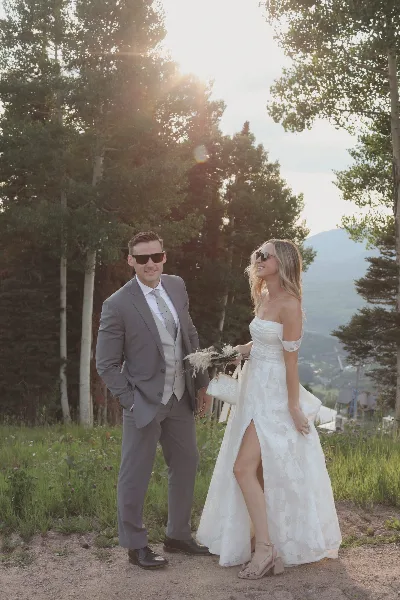 A bride and groom standing outdoors in a mountainous landscape, wearing formal wedding attire.