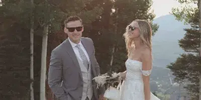 A bride and groom standing outdoors in a mountainous landscape, wearing formal wedding attire.