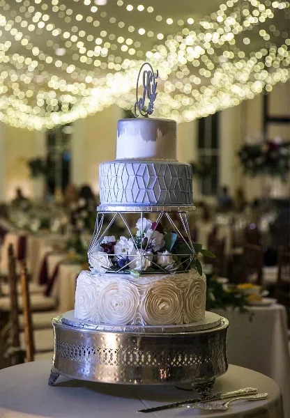A three-tiered white wedding cake with geometric and floral decorations, topped with a monogram, displayed on a silver stand in a banquet hall.