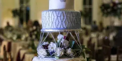 A three-tiered white wedding cake with geometric and floral decorations, topped with a monogram, displayed on a silver stand in a banquet hall.